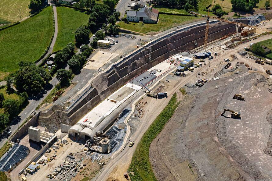 Luftaufnahme des fortgeschrittenen Tunnelbaus der Ortsumgehung Hosingen mit zwei sichtbaren Tunnelportalen, Baucontainern, Zufahrtsbauwerk und stabilisiertem Hang