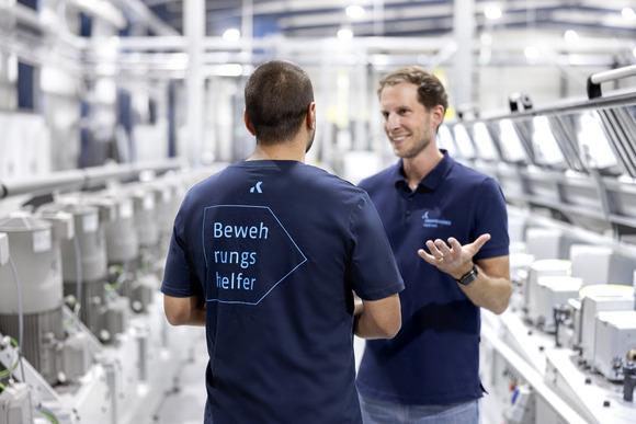 Zwei M&auml;nner in Arbeitskleidung stehen in einer Produktionshalle, einer spricht, der andere h&ouml;rt aufmerksam zu.