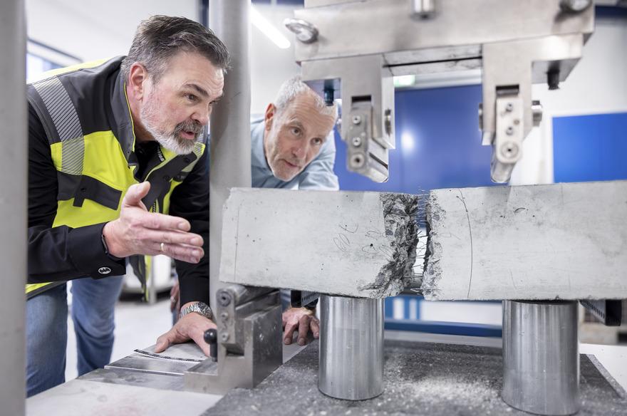 Two men observe a metal specimen being tested for pressure in a laboratory setting, examining the results closely.